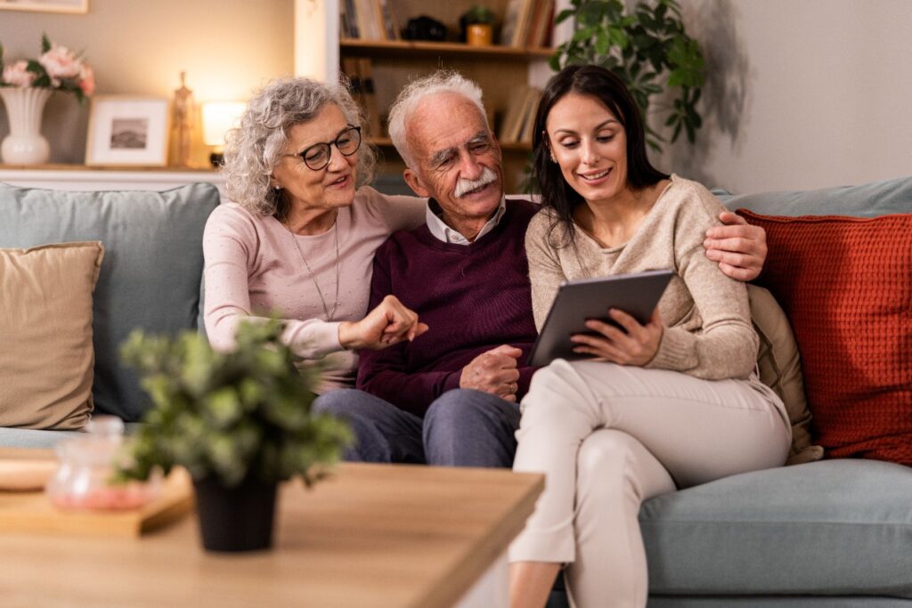 A senior couple and their adult daughter sit together on a couch, smiling as they look at a tablet in a warm, comfortable living room — a perfect snapshot of independence in retirement living.