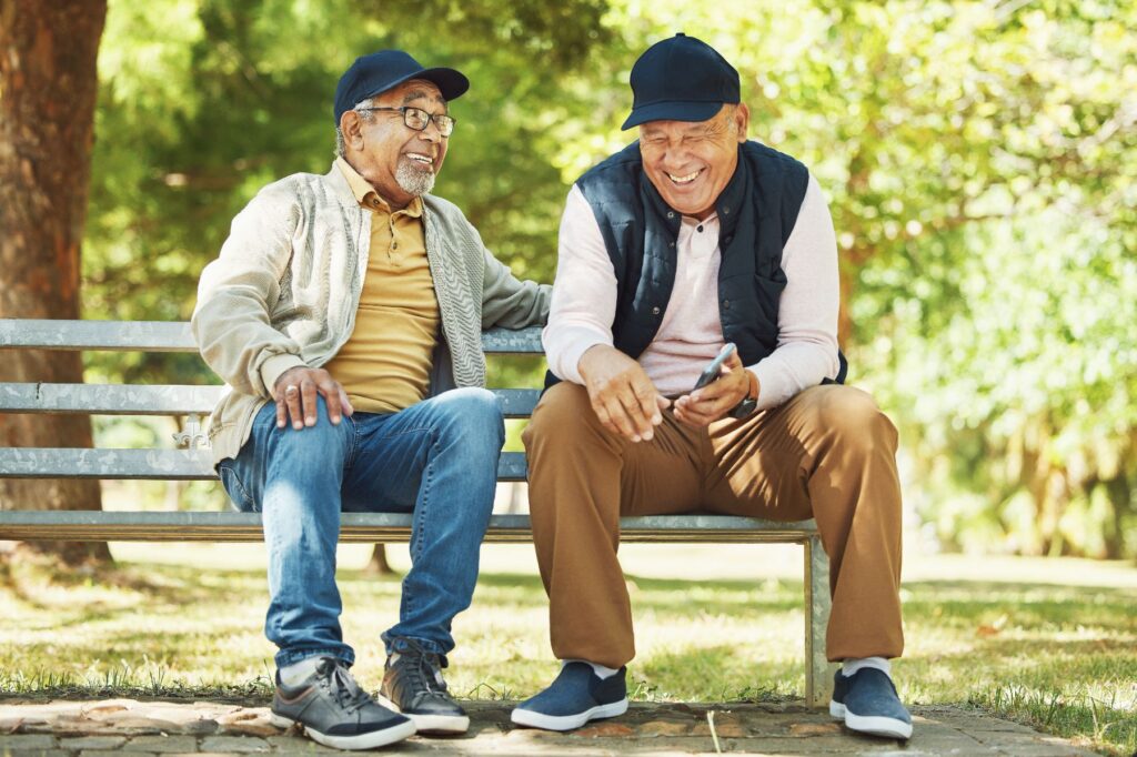 Two older men share a laugh on a sunny park bench surrounded by trees, enjoying the kind of outdoor connection that highlights the benefits of nature for seniors and the welcoming spirit of senior living in Brevard, NC.