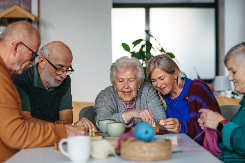 Five older adults lean in together over a shared activity at a community table, laughing and engaging side by side in the kind of social activities for seniors that make everyday life feel genuinely full.