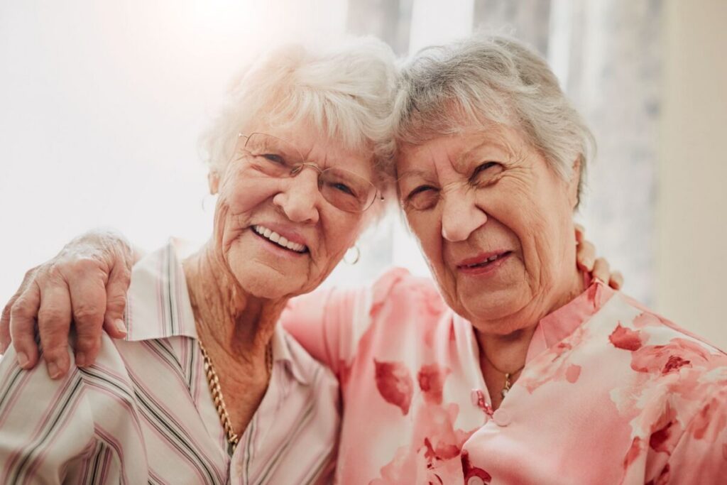 Two older women lean their heads together, smiling warmly with arms wrapped around each other's shoulders — a close, joyful friendship that reflects the benefits of small senior living communities, where residents build genuine, familiar bonds with the people around them.